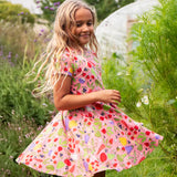 Child wearing Frugi's 100% organic cotton spring skater dress in pink with a repeating fruit and veg print with grass and flowers in the background