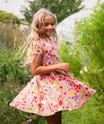 Child wearing Frugi's 100% organic cotton spring skater dress in pink with a repeating fruit and veg print with grass and flowers in the background