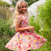 Child wearing Frugi's 100% organic cotton spring skater dress in pink with a repeating fruit and veg print with grass and flowers in the background
