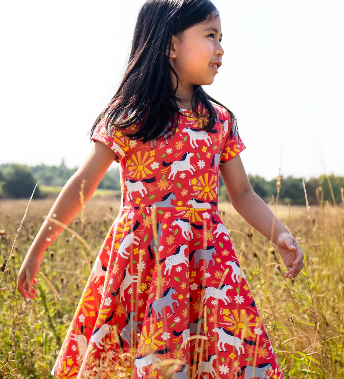 Child wearing Frugi 100% spring skater dress in coral pink with repeating prints of grey and white horses in a grassy field