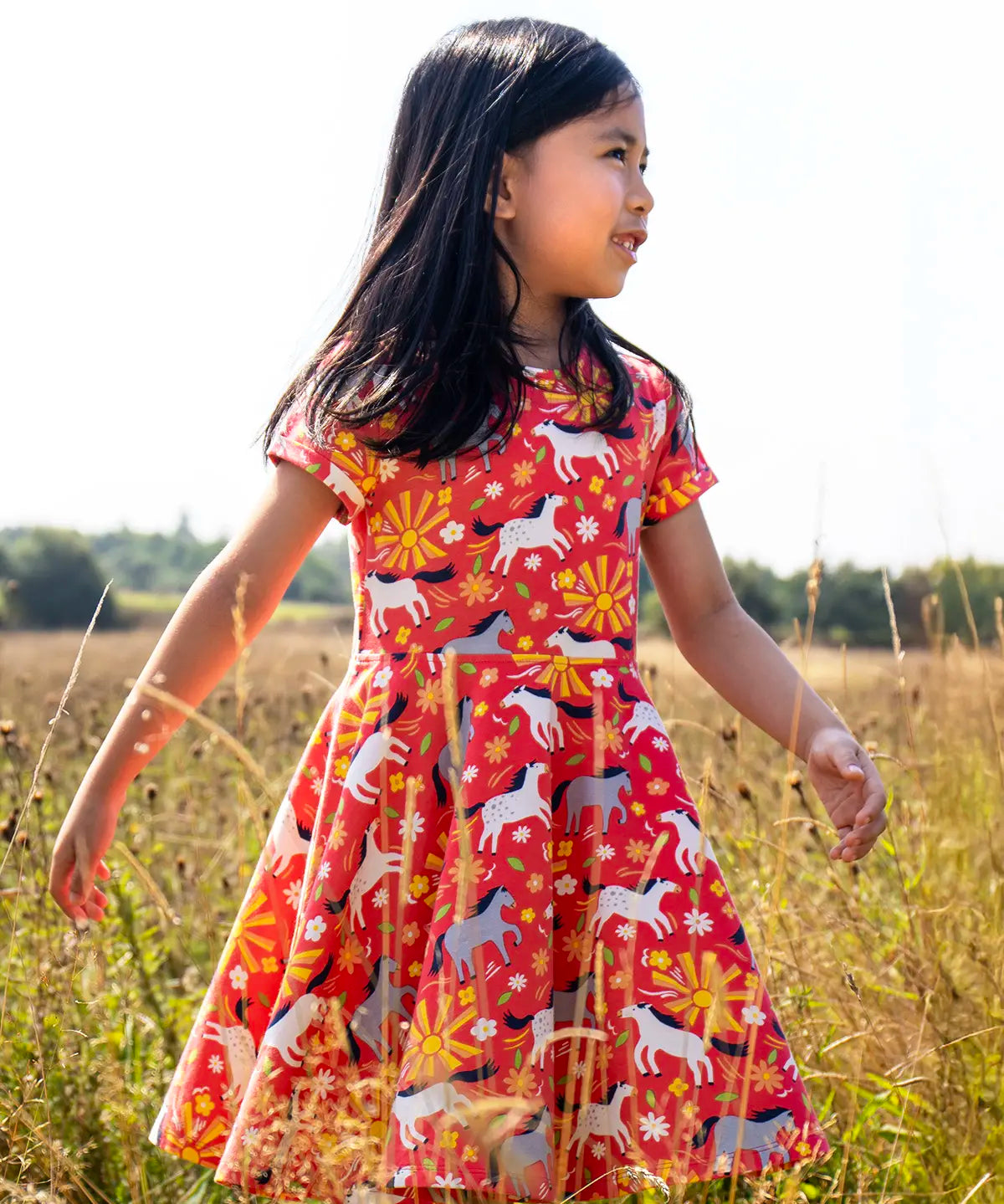 Child wearing Frugi 100% spring skater dress in coral pink with repeating prints of grey and white horses in a grassy field