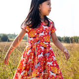 Child wearing Frugi 100% spring skater dress in coral pink with repeating prints of grey and white horses in a grassy field