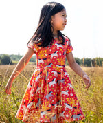 Child wearing Frugi 100% spring skater dress in coral pink with repeating prints of grey and white horses in a grassy field