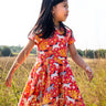Child wearing Frugi 100% spring skater dress in coral pink with repeating prints of grey and white horses in a grassy field