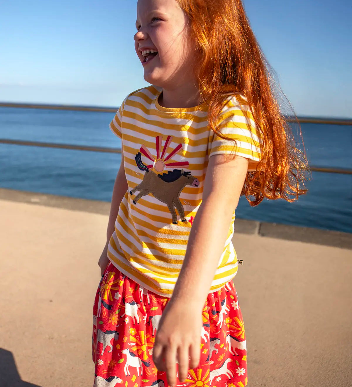 Child wearing Frugi 100% organic cotton spring skort in coral pink with a repeating white and grey pony print with the ocean in the background