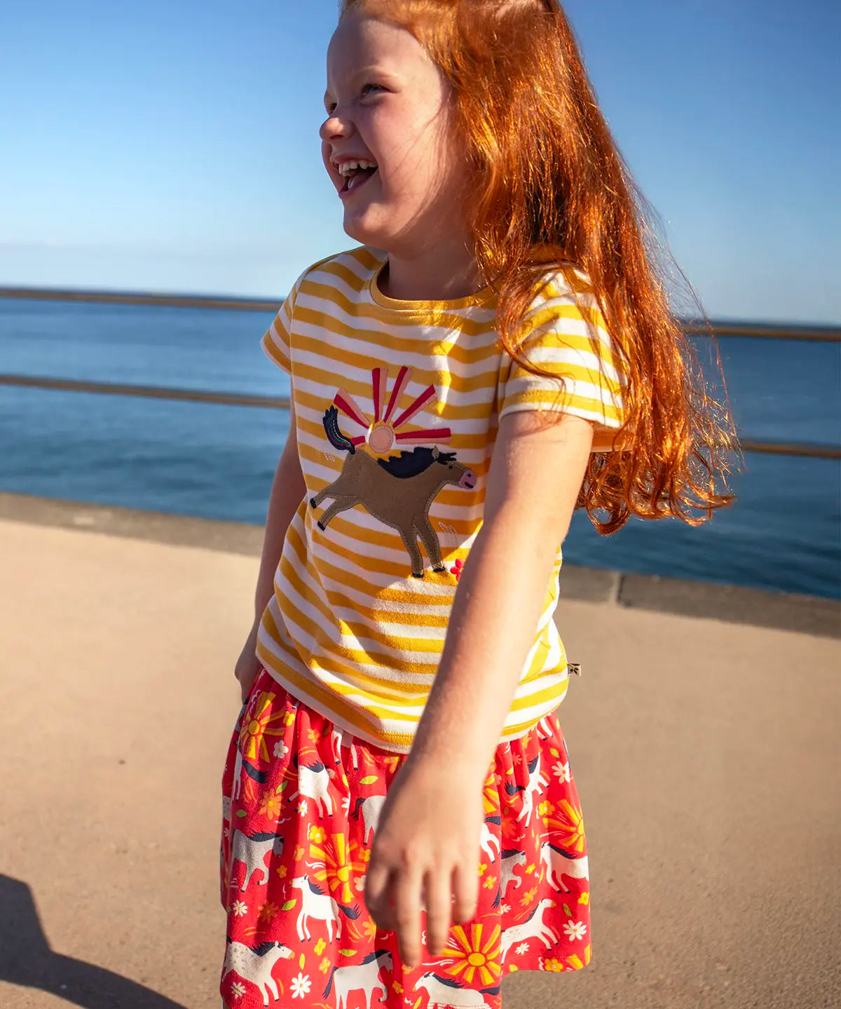 Child wearing Frugi 100% organic cotton spring skort in coral pink with a repeating white and grey pony print with the ocean in the background
