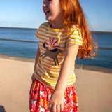 Child wearing Frugi 100% organic cotton spring skort in coral pink with a repeating white and grey pony print with the ocean in the background