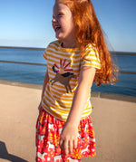 Child wearing Frugi 100% organic cotton spring skort in coral pink with a repeating white and grey pony print with the ocean in the background