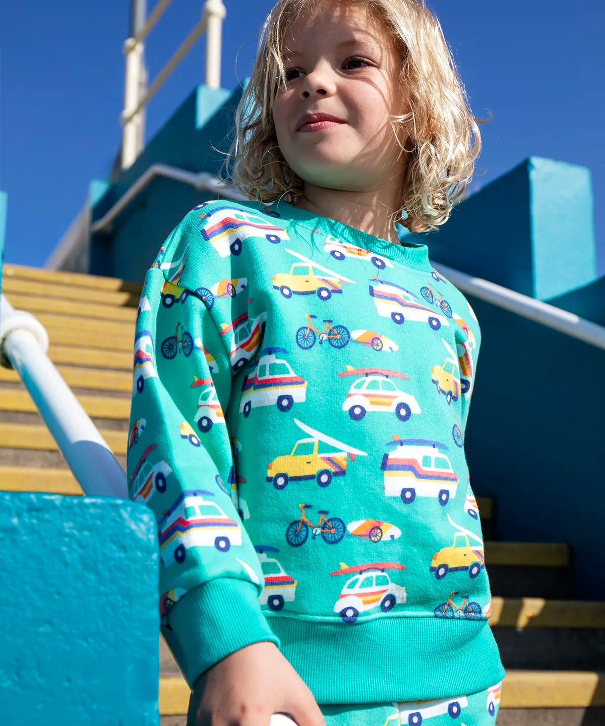 Child wearing Frugi 100% organic cotton sweatshirt in light green with repeating beach inspired vehicles. Child on concrete stairs