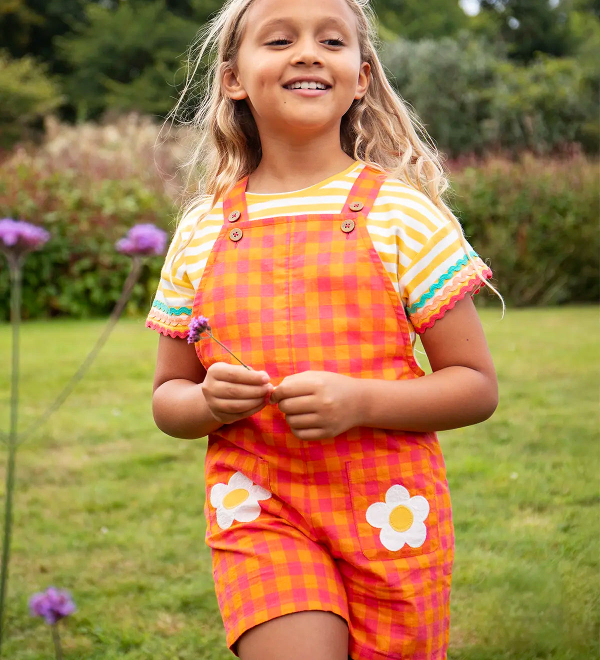 Child wearing Frugi 100% organic cotton Thea yellow and white striped t-shirt with a ric rac decorative trim with dungarees in a grassy field