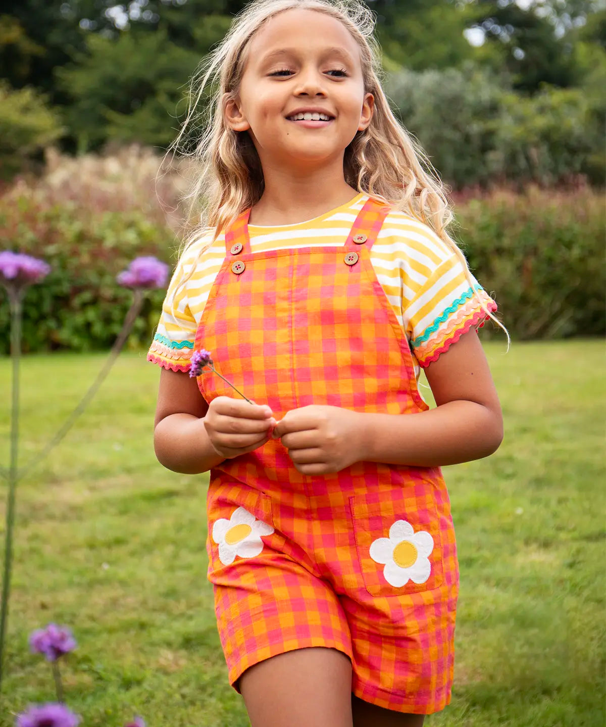 Child wearing Frugi 100% organic cotton Thea yellow and white striped t-shirt with a ric rac decorative trim with dungarees in a grassy field