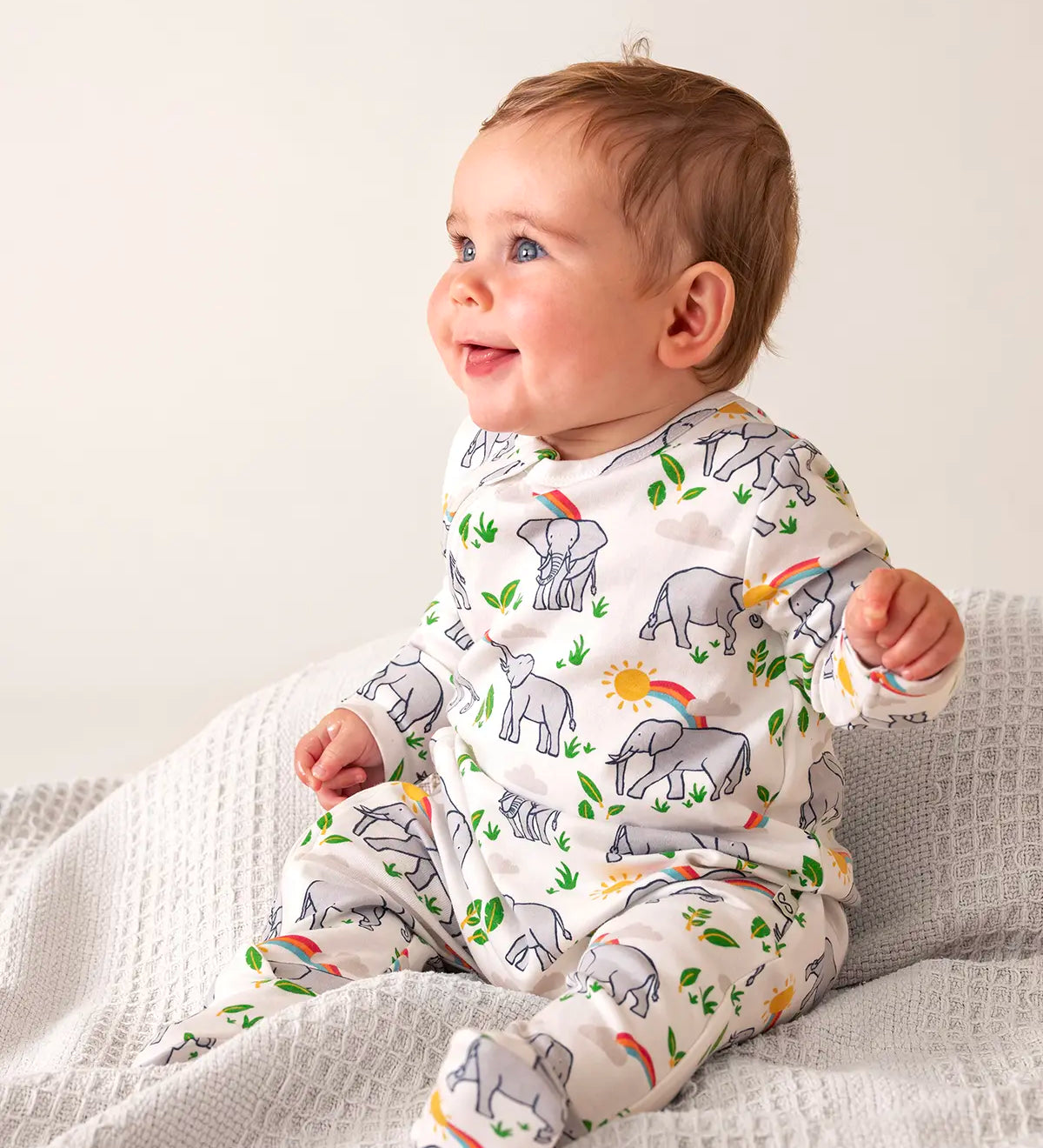 Baby wearing Frugi's 100% organic cotton kimono style babygrow in white with a repeating elephant and rainbow print on a grey blanket