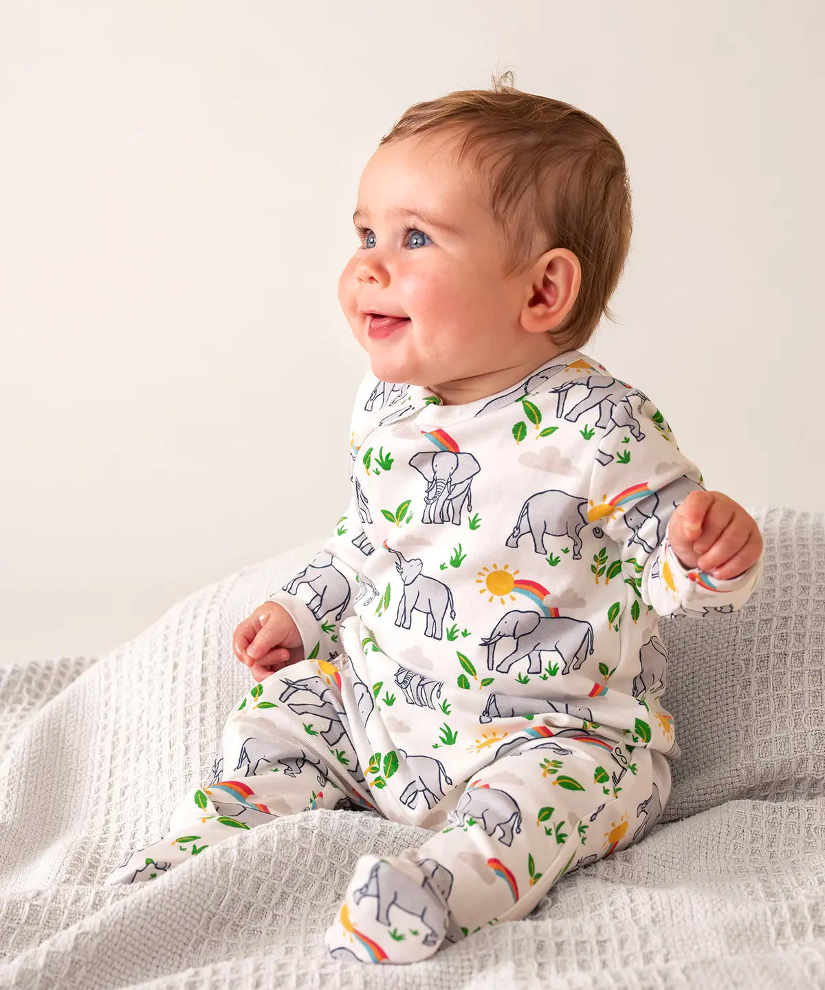 Baby wearing Frugi's 100% organic cotton kimono style babygrow in white with a repeating elephant and rainbow print on a grey blanket