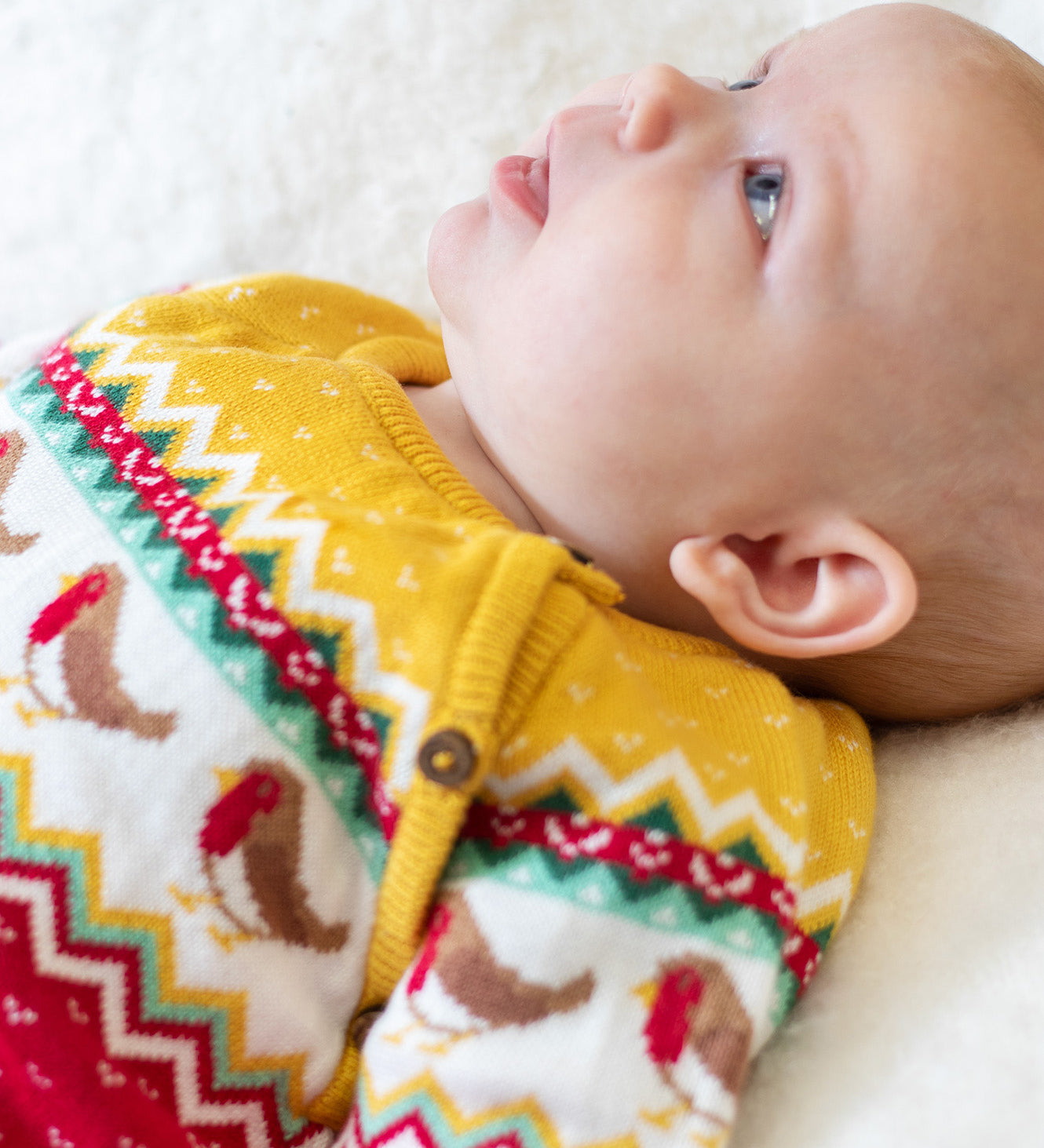 A baby wearing the Frugi lumi knitted festive romper fair isle available at Babipur. A GOTS organic cotton soft knitted all in one in a fairisle design with robins. Features button fasteners running down from the shoulder.