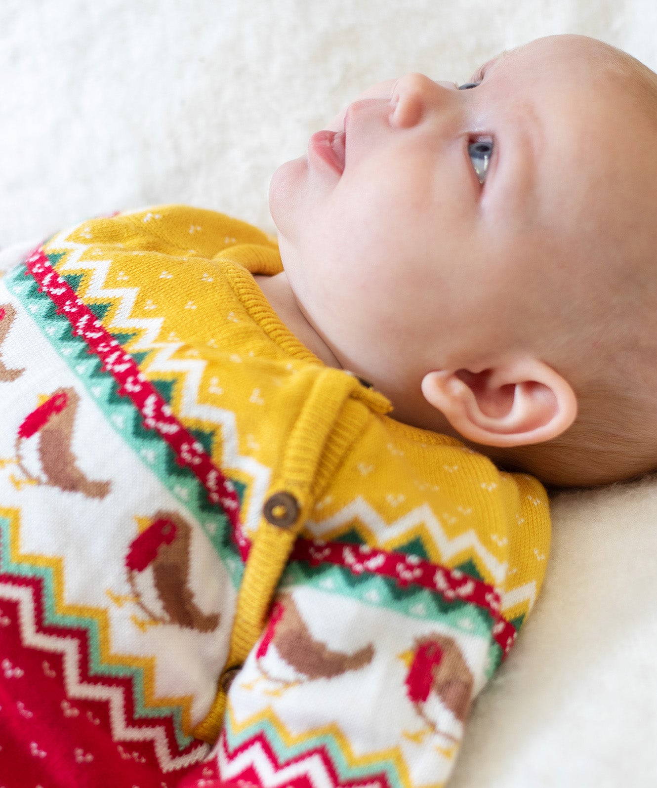 A baby wearing the Frugi lumi knitted festive romper fair isle available at Babipur. A GOTS organic cotton soft knitted all in one in a fairisle design with robins. Features button fasteners running down from the shoulder.