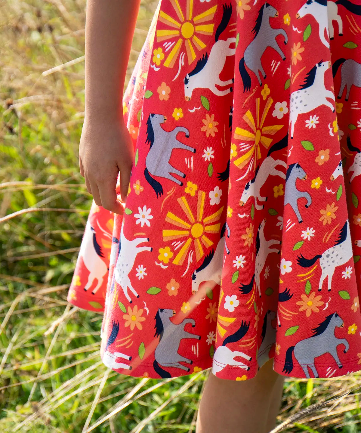 Close up of child wearing Child wearing Frugi 100% spring skater dress in coral pink with repeating horses and  showing the frill on the hem  