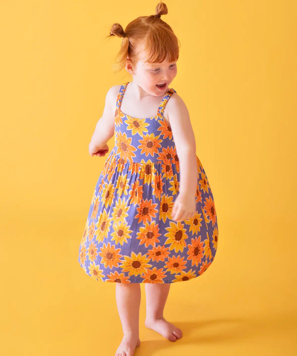 Child wearing Frugi 100% organic cotton Sophie summer sleeveless dress in purple with a repeating sunflower print against a orange background