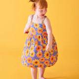 Child wearing Frugi 100% organic cotton Sophie summer sleeveless dress in purple with a repeating sunflower print against a orange background