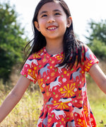 Child wearing Frugi 100% spring skater dress in coral pink with repeating prints of grey and white horses with trees in the background