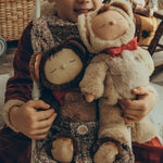 Olli Ella Cozy Dinkum Dolls Teddy Twiggy and Teddy Crumble together, sat in an Olli Ella Stroller with a child sat behind holding onto the back of the stroller. The child is wearing a red Santa outfit and is smiling down at Teddy Crumble