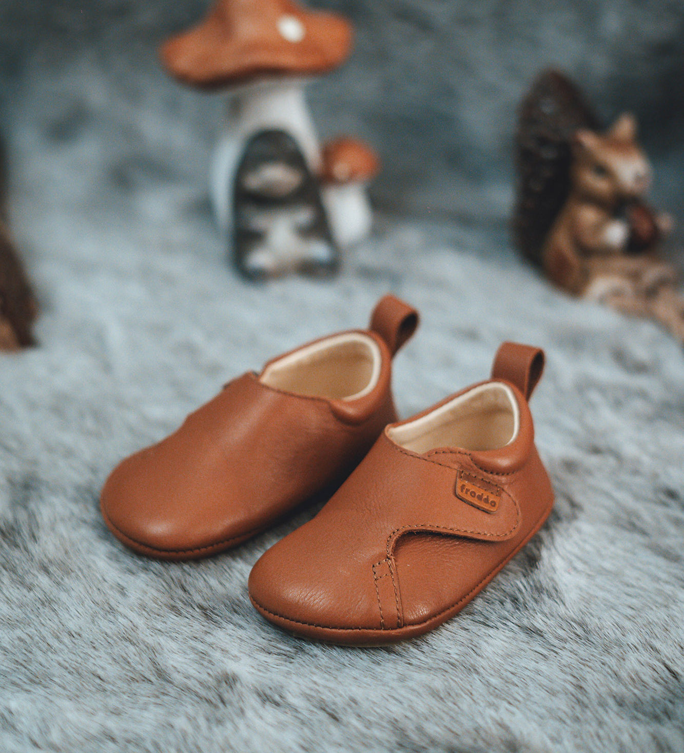 Froddo barefoot walker baby shoe in tan with a autumn display in the background. A light brown natural leather shoe with a pull tab on the back for easy fitting, a velcro fastener, a grippy sole and available at Babipur.