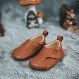 Froddo barefoot walker baby shoe in tan with a autumn display in the background. A light brown natural leather shoe with a pull tab on the back for easy fitting, a velcro fastener, a grippy sole and available at Babipur.