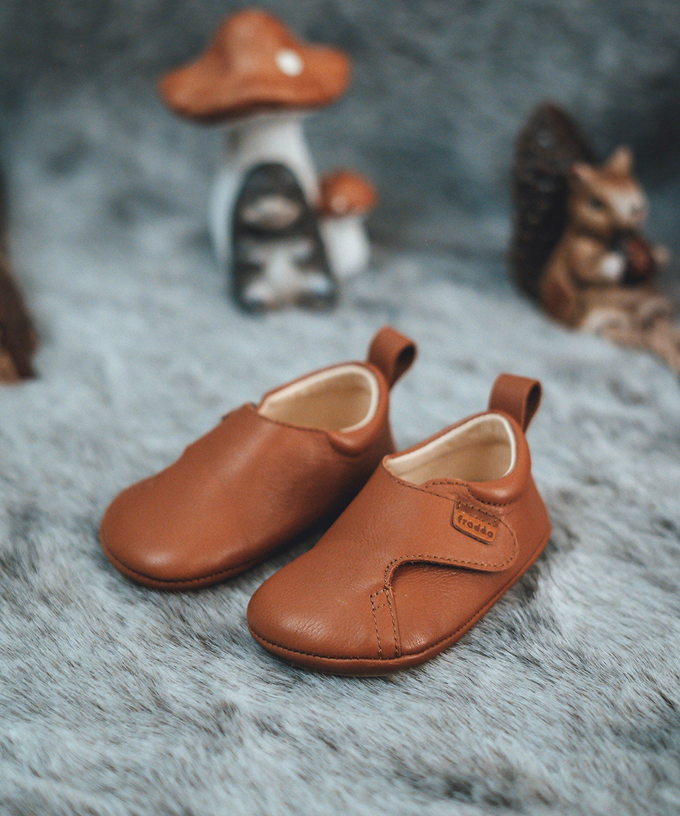 Froddo barefoot walker baby shoe in tan with a autumn display in the background. A light brown natural leather shoe with a pull tab on the back for easy fitting, a velcro fastener, a grippy sole and available at Babipur.