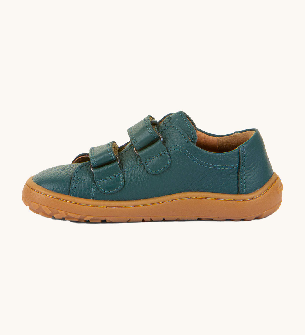 A side image of the Froddo barefoot base children's shoe in petroleum available at Babipur. A dark turquoise natural leather low profile shoe showing the 2 velcro fasteners looped through and the brown grippy sole