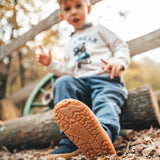 Child sitting on a log and wearing Froddo’s Barefoot Kids F-Motion TEX Shoes in dark blue showing close up of the flexible brown rubber sole tread