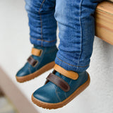 A child wearing the Froddo barefoot first step baby shoe in dark denim available at Babipur. A dark blue natural leather shoe with a 2 velcro fastener in dark yellow and brown for a secure fit. Features a light brown grippy sole