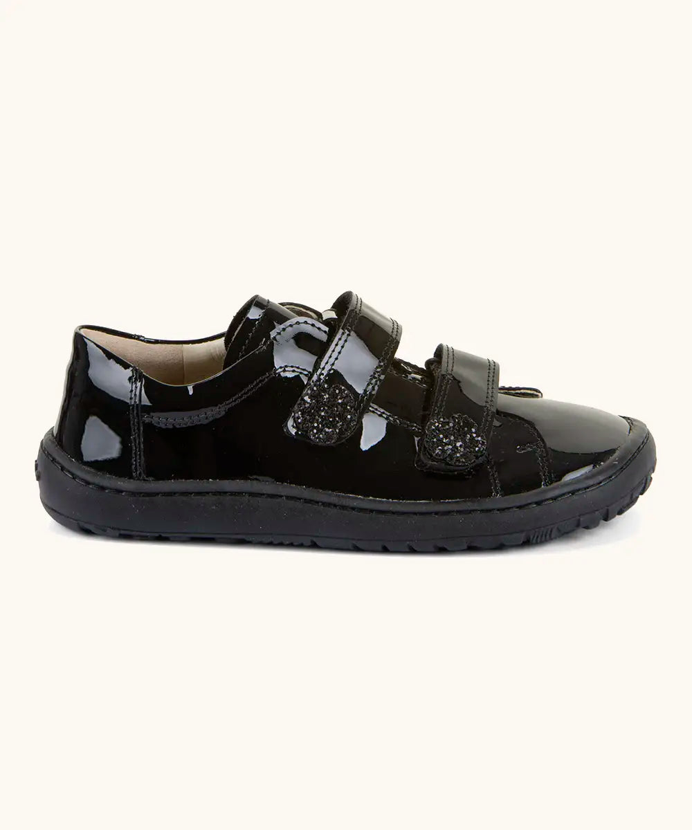 Side of Froddo barefoot kids school shoe in black patent. A low profile shoe with 2 front velcro straps with a decoration on the end