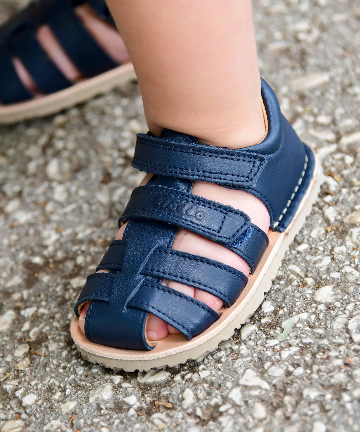 Small child wearing Froddo's barefoot natural leather sandal in dark blue standing on a concrete surface