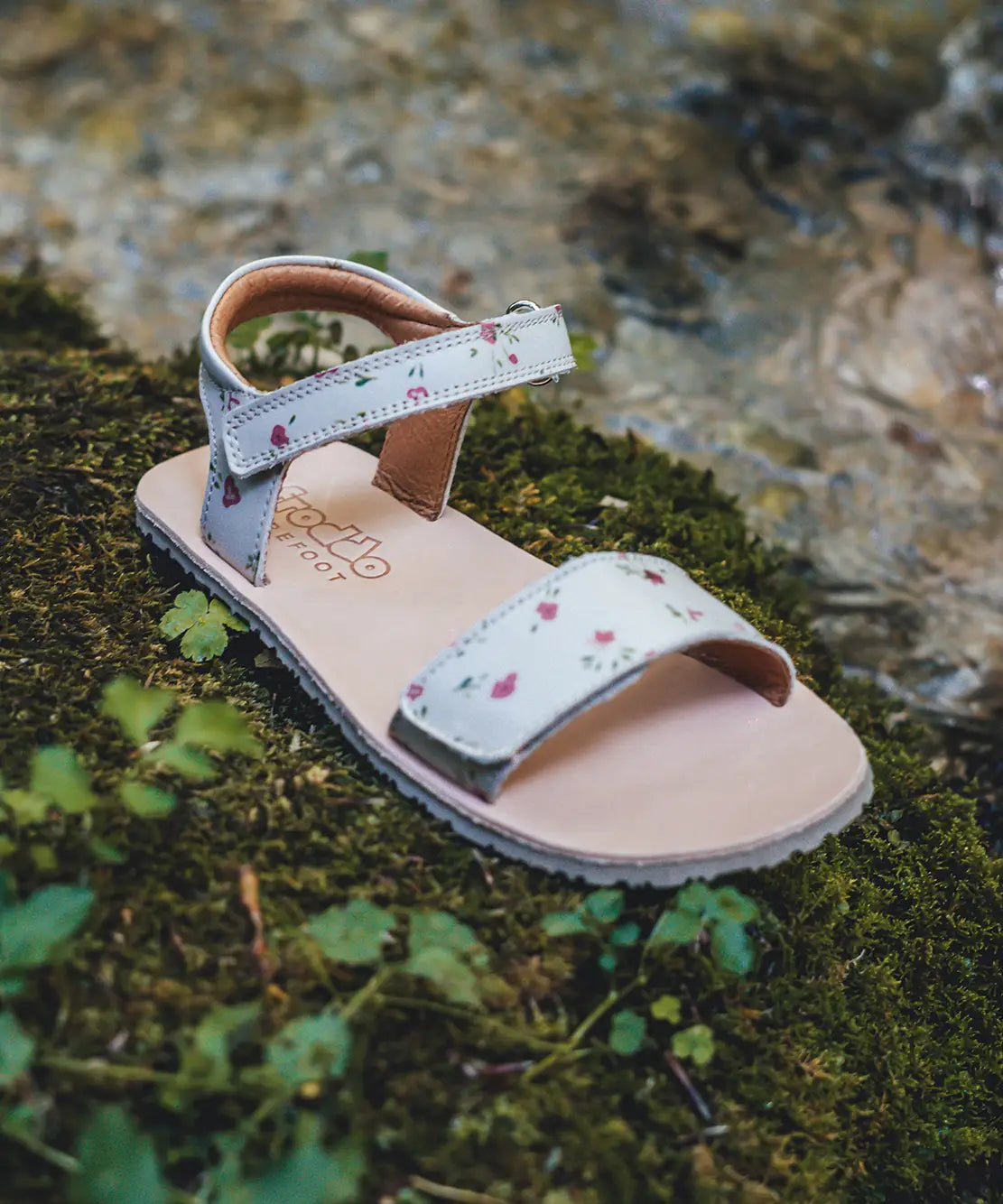 A Froddo's barefoot kids natural leather Lia sandal in pink with flowers standing on green moss