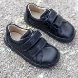 Pair of Froddo barefoot natural leather kids school shoes in black with velcro fasteners on a concrete surface