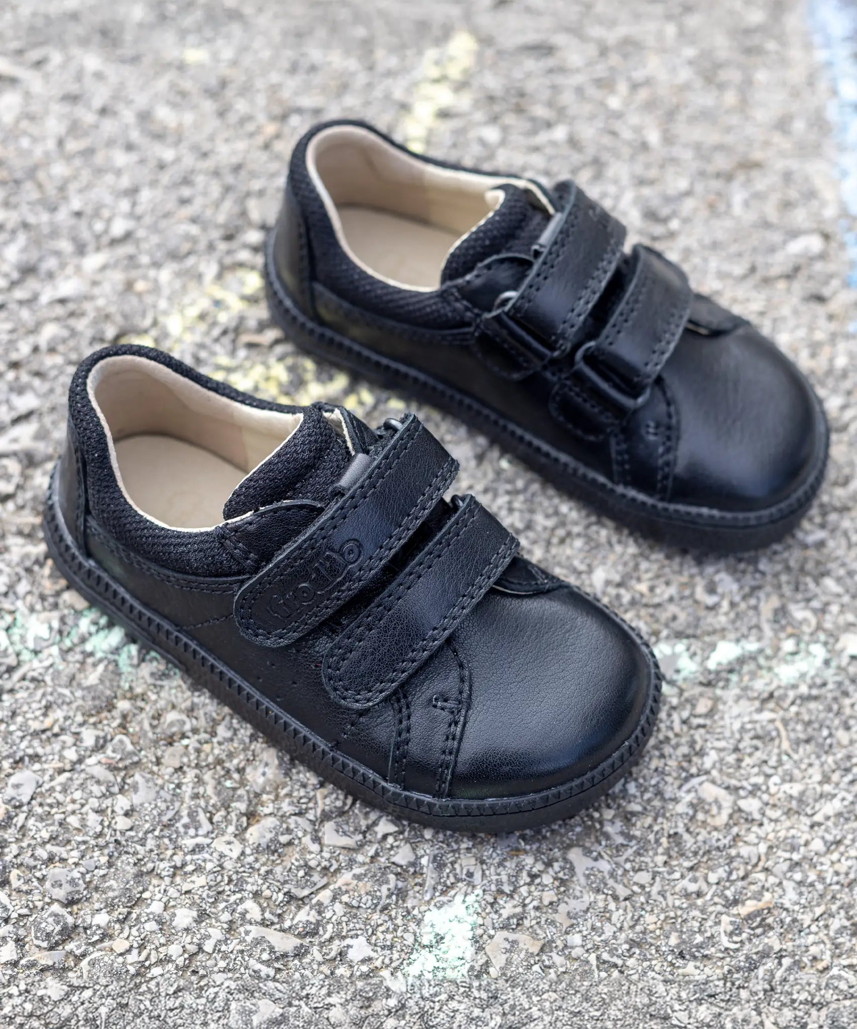 Pair of Froddo barefoot natural leather kids school shoes in black with velcro fasteners on a concrete surface
