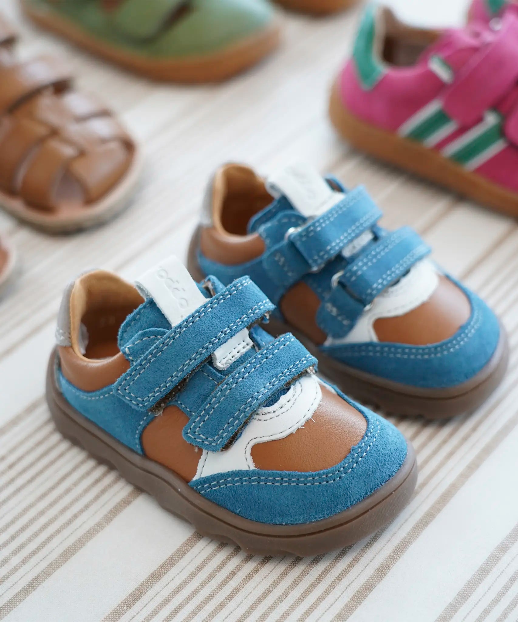 Froddo barefoot zeru Spring kids shoes in blue brown and white standing on a beige striped floor next to other trainers and sandals