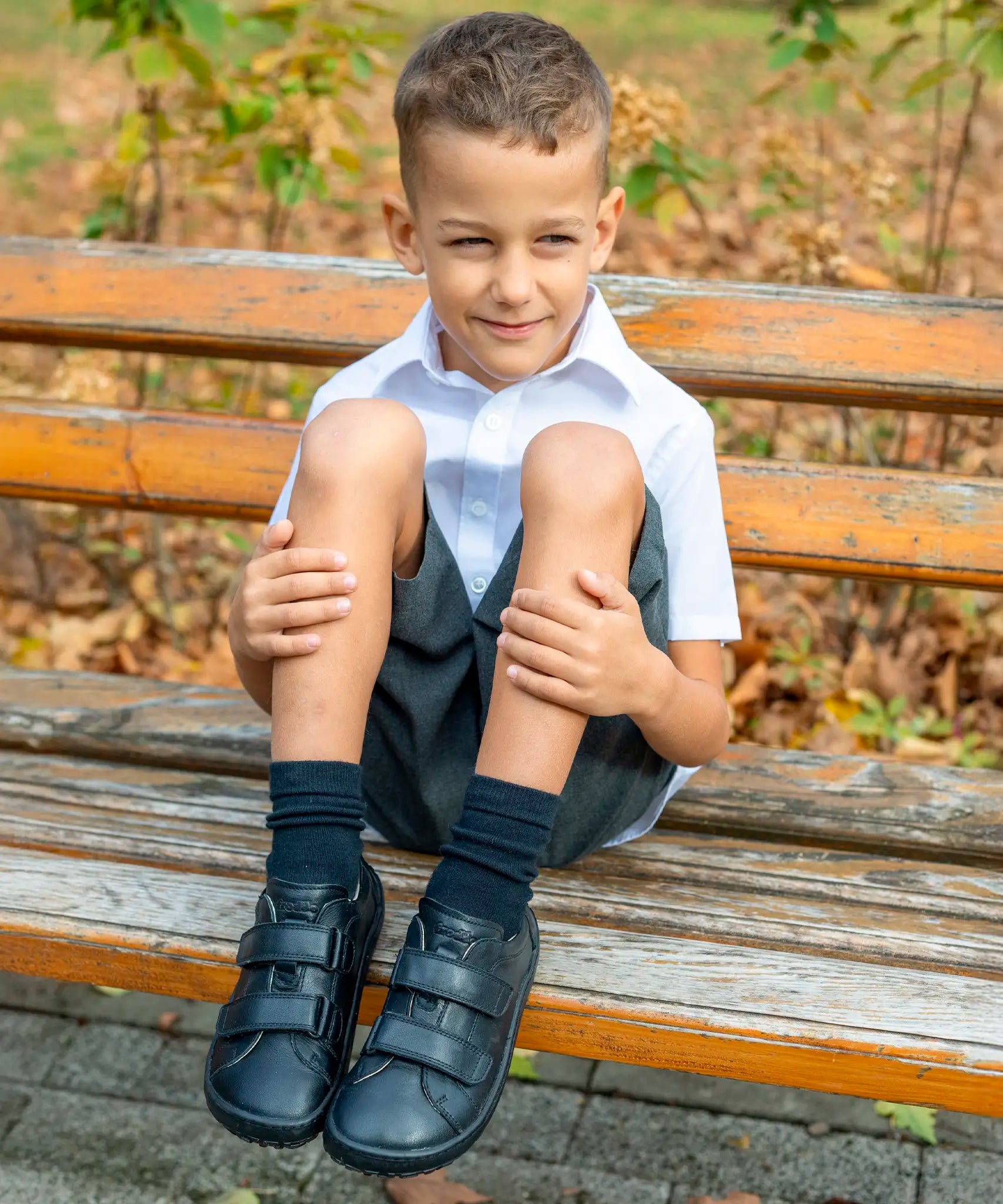 Child sitting on a wooden bench wearing Froddo's natural black leather barefoot school shoes showing front velcro fasteners