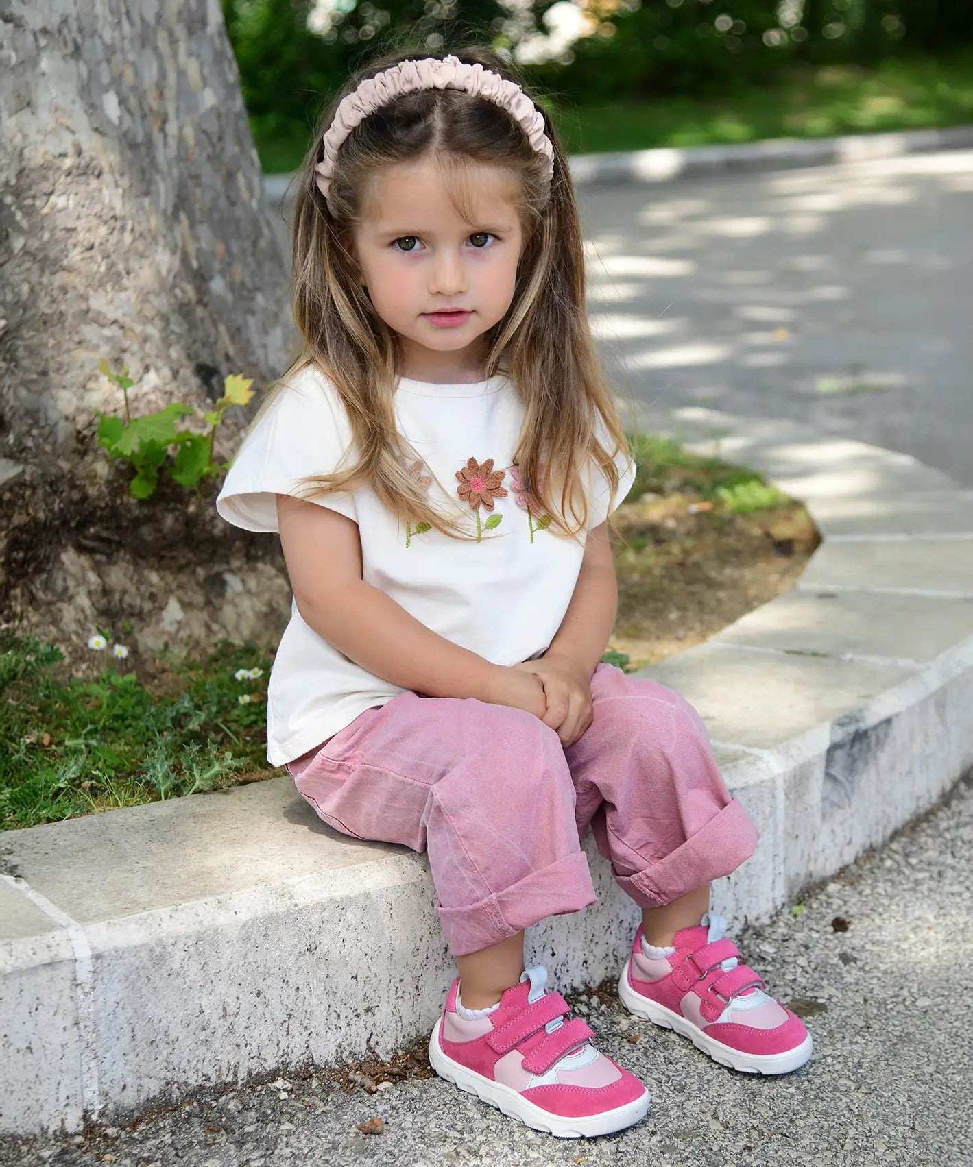 Small child wearing Froddo's barefoot zeru Spring kids shoes in light and dark pink. Sitting on a stone surface with trees and plants in the background