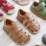 Pair of Froddo barefoot kids flexy sandals in tan standing on a beige striped floor showing brand name on veclro strap. Sandals standing next to other shoes