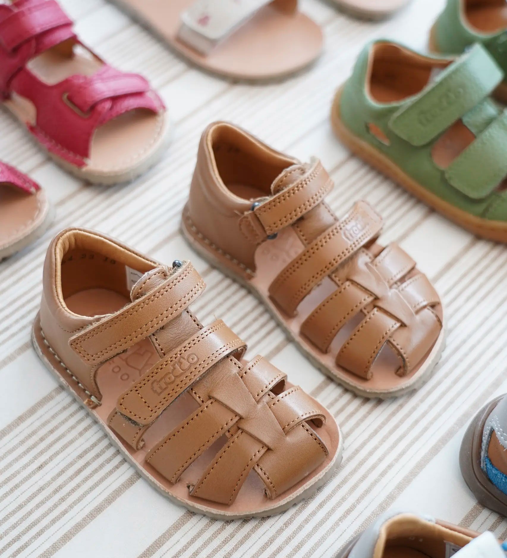 Pair of Froddo barefoot kids flexy sandals in tan standing on a beige striped floor showing brand name on veclro strap. Sandals standing next to other shoes