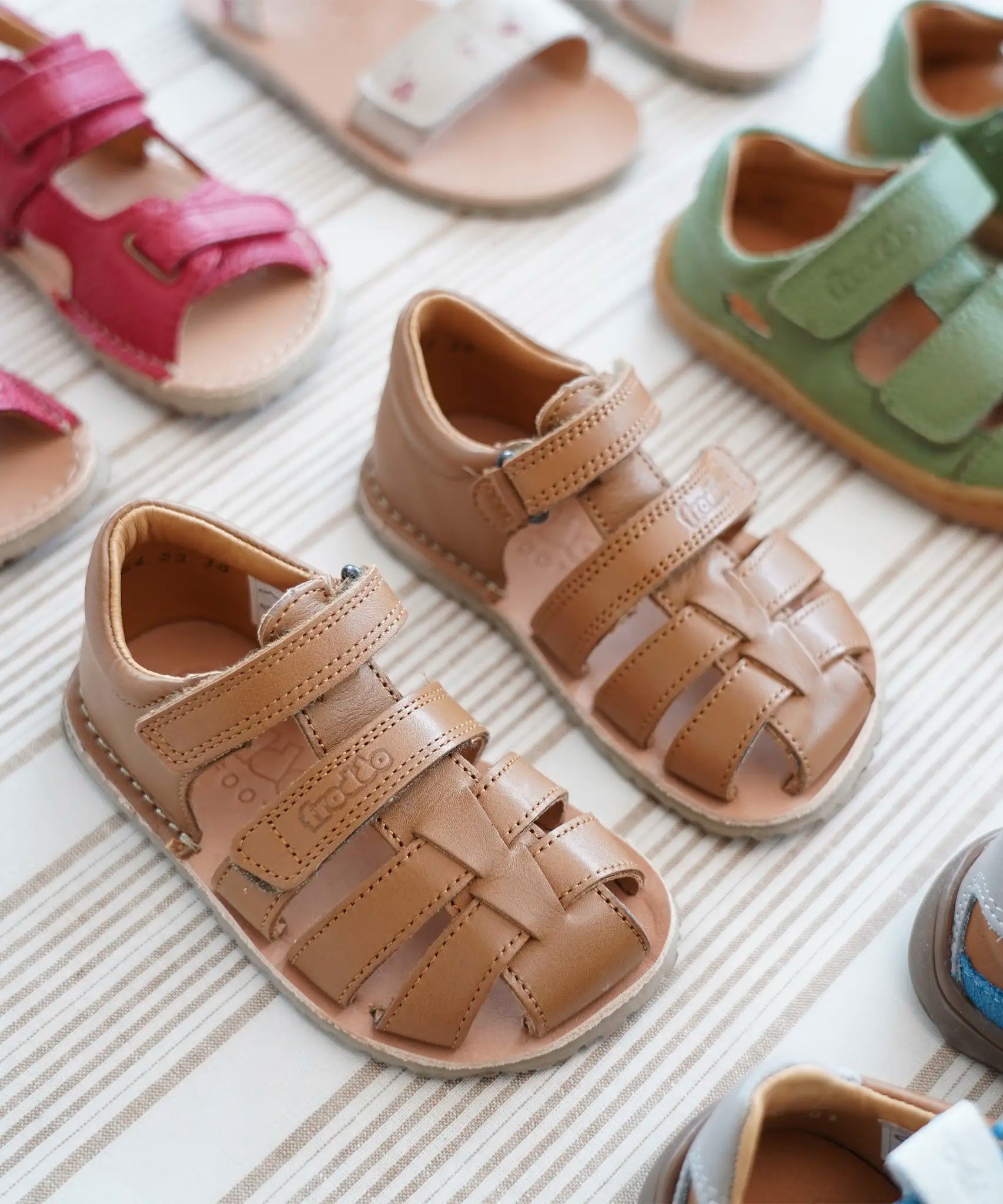 Pair of Froddo barefoot kids flexy sandals in tan standing on a beige striped floor showing brand name on veclro strap. Sandals standing next to other shoes