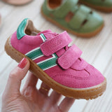 Adult hand holding a Froddo barefoot f-motion kids shoe in bright pink with green stripe design on the side showing brand name on velcro strap