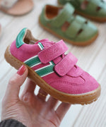 Adult hand holding a Froddo barefoot f-motion kids shoe in bright pink with green stripe design on the side showing brand name on velcro strap