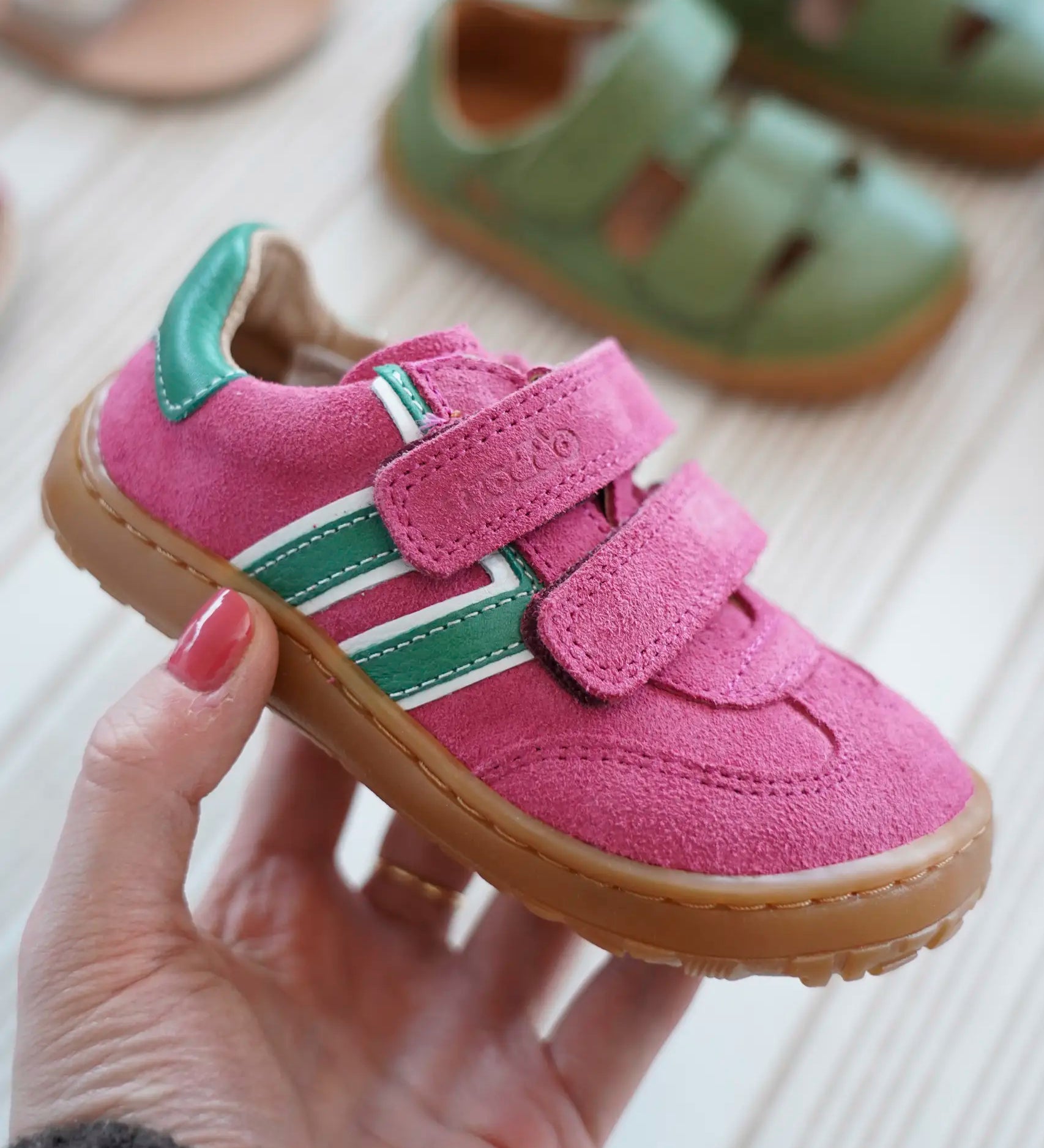 Adult hand holding a Froddo barefoot f-motion kids shoe in bright pink with green stripe design on the side showing brand name on velcro strap