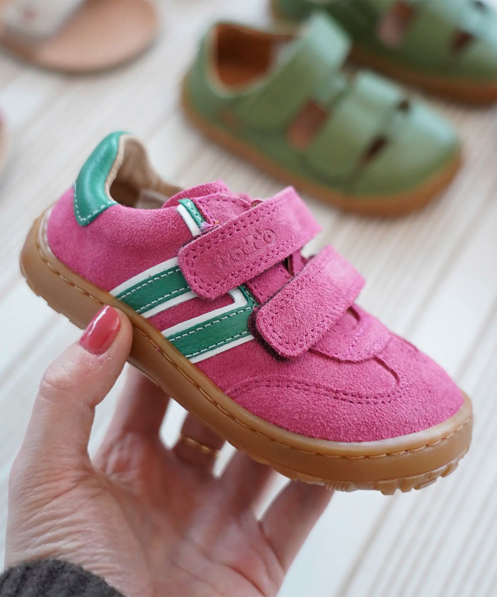 Adult hand holding a Froddo barefoot f-motion kids shoe in bright pink with green stripe design on the side showing brand name on velcro strap