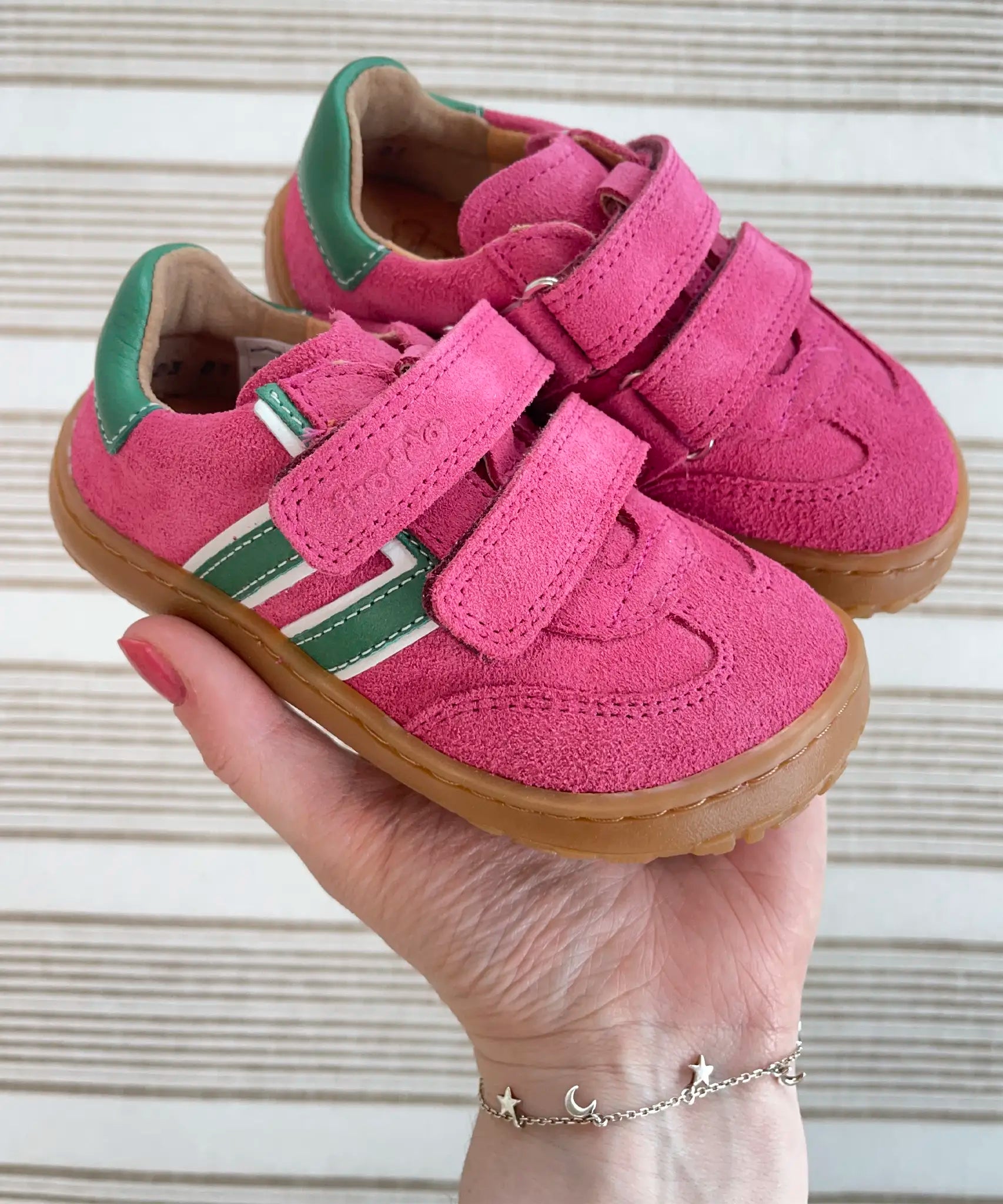 Adult holding Froddo's barefoot f-motion kids trainers in bright pink with green stripe design showing brand name on velcro strap