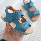 Adult hand holding Froddo's light blue barefoot kids natural leather sandal showing brand name on front velcro strap and padded ankle support
