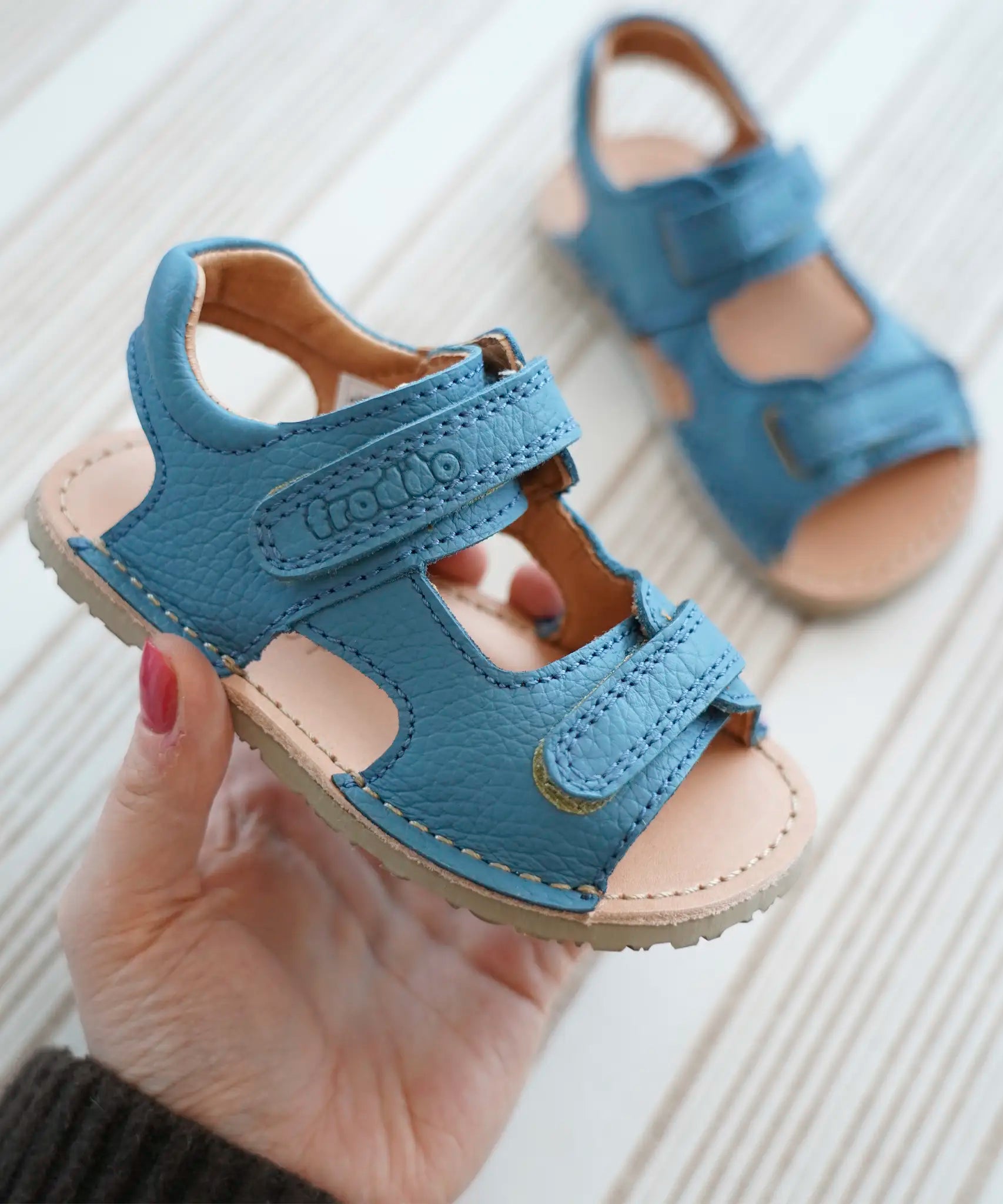 Adult hand holding Froddo's light blue barefoot kids natural leather sandal showing brand name on front velcro strap and padded ankle support