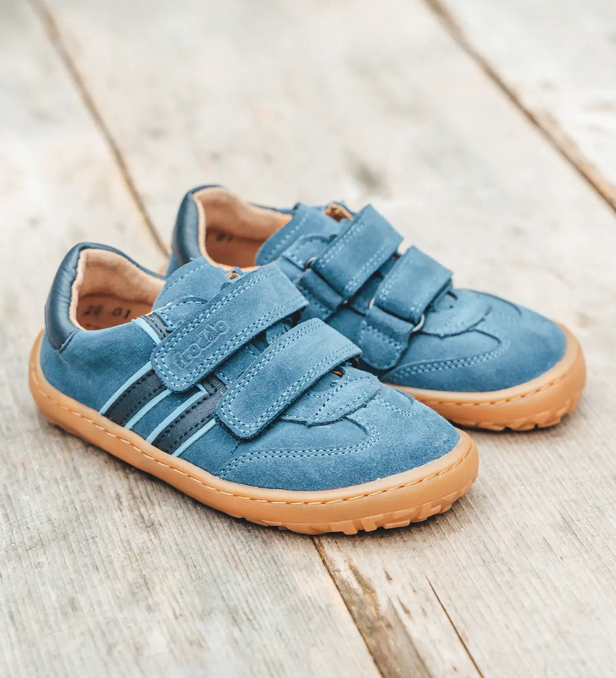 Pair of Froddo barefoot f-motion kids suede shoes in denim blue on decking. Sports trainer has a wide, foot shaped toe box and zero heel drop