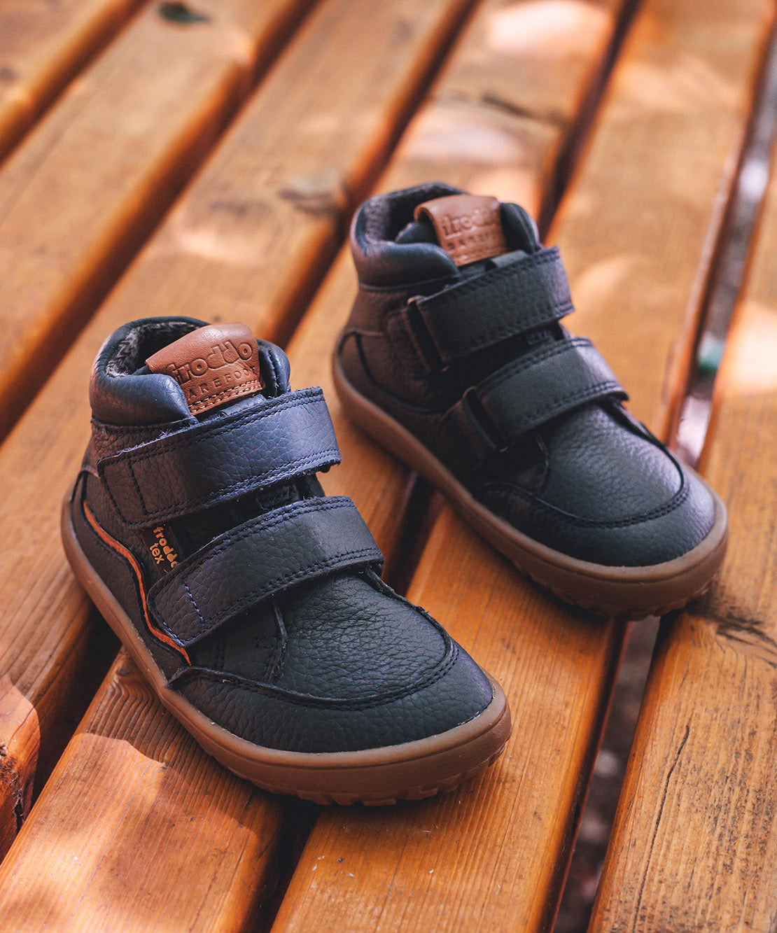 Froddo barefoot tex kids autumn shoes in navy standing on a wooden surface and available at Babipur. Soft ankle high shoes in navy with a light brown rubber sole. Features two velcro straps for a secure fit 